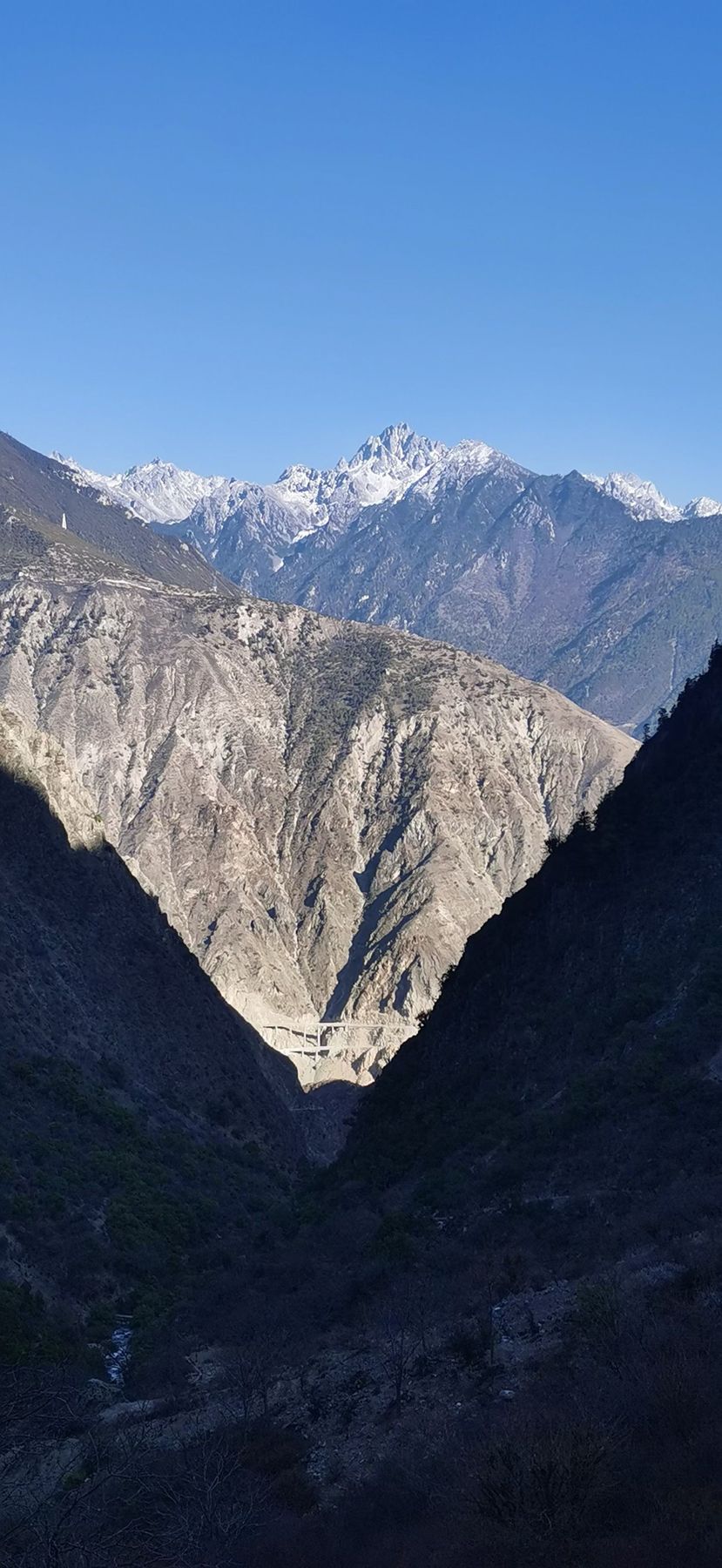 A snow-capped peak above a deep V-shaped valley with a road bridge below