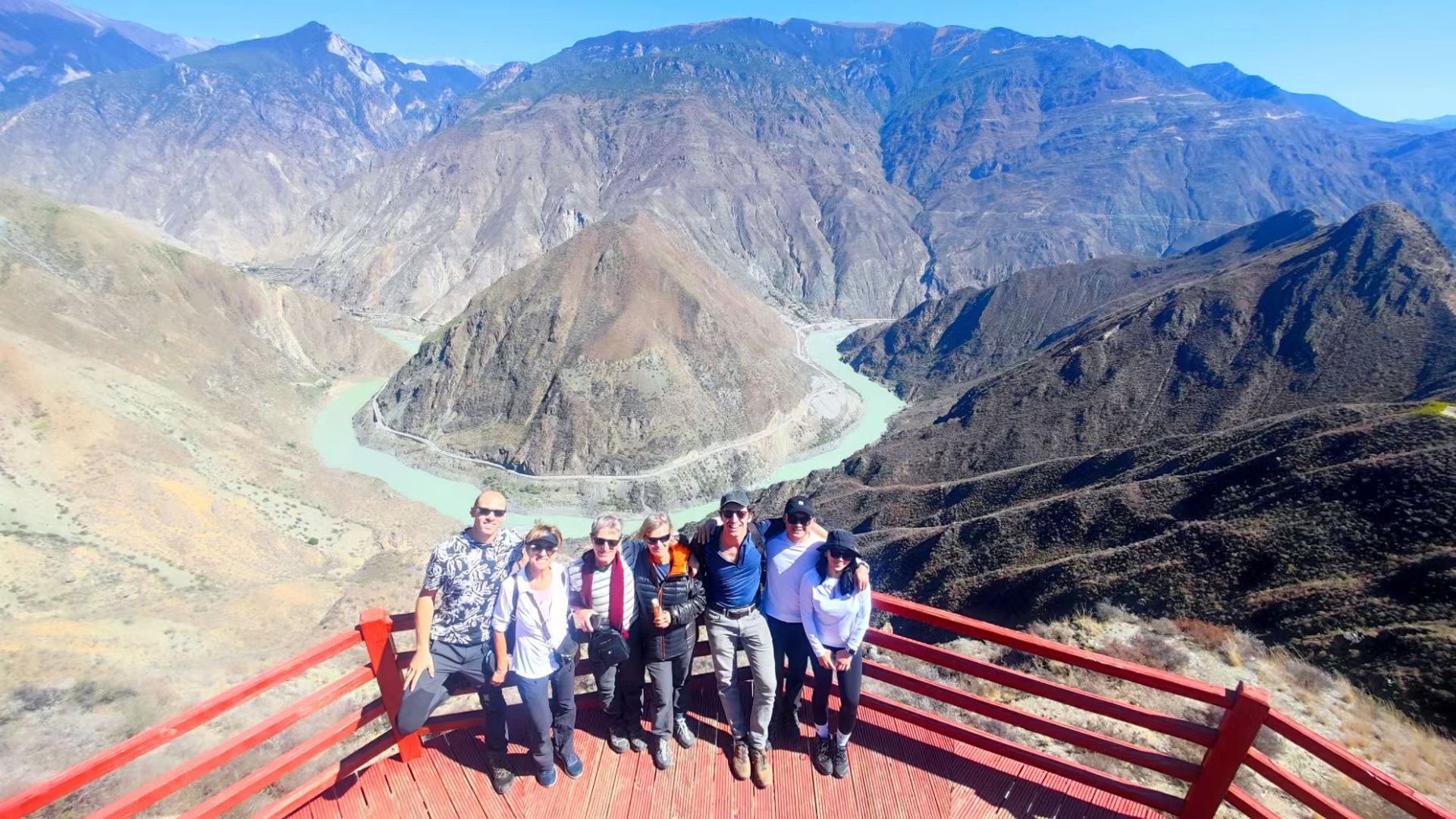 Seven hikers on a red viewing platform above a tight bend of the pale green Jinsha River