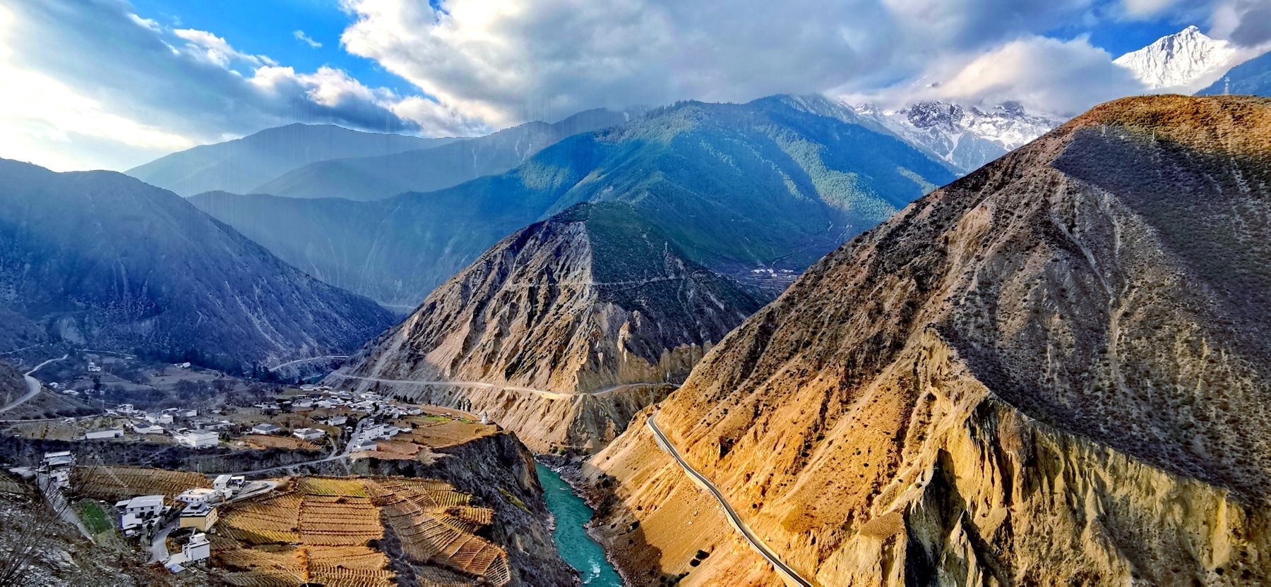 A deep valley with the turquoise Mekong, a village on terraces and a snow peak in the clouds