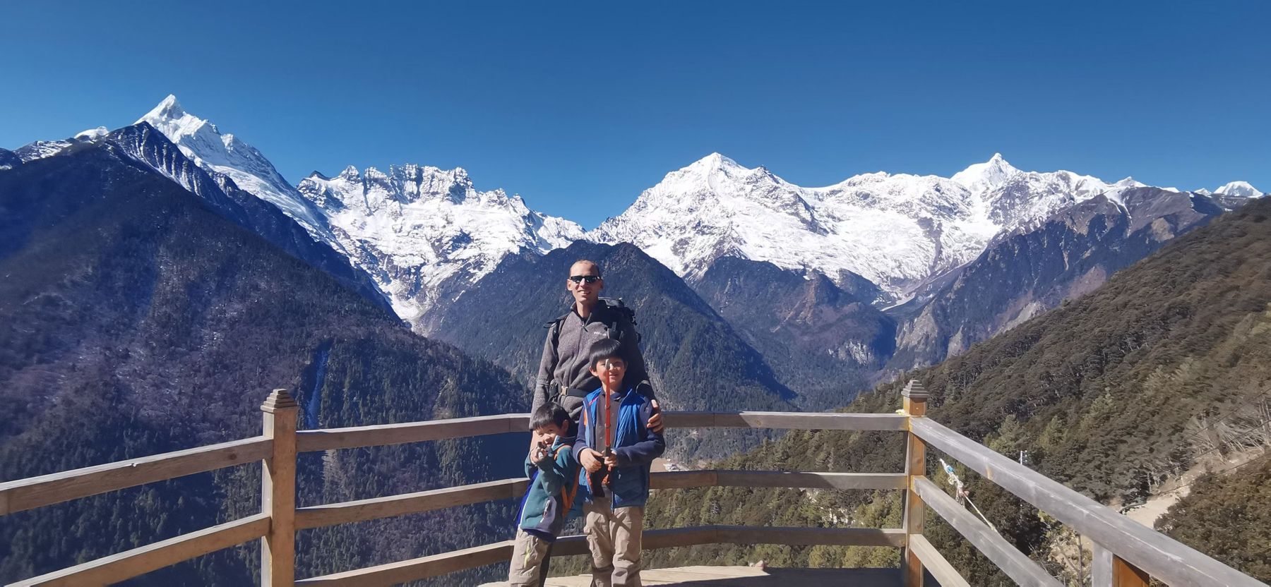 A man and a boy at a wooden railing on a pass, facing a range of snow peaks