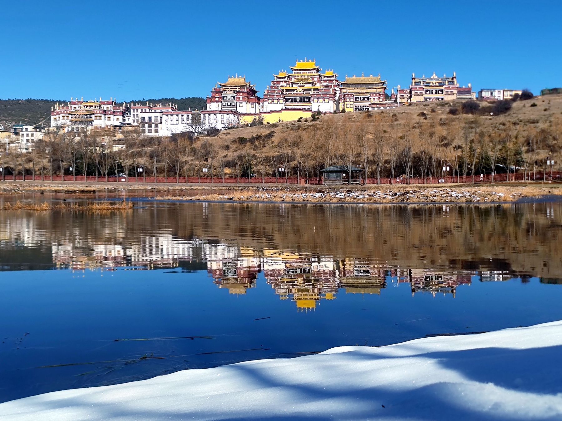A golden-roofed monastery reflected in a lake, with snow in the foreground